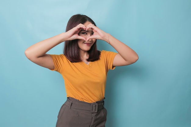 Attractive young Asian woman feels happy and romantic shapes heart gesture expresses tender feelings wears casual yellow tshirt against blue background People affection and care concept