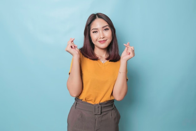 Attractive young Asian woman feels happy and romantic shapes heart gesture expresses tender feelings wears casual yellow tshirt against blue background People affection and care concept