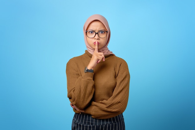 Attractive young Asian woman finger on lips and making silent gesture on blue background