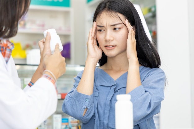 Attractive young asian woman having a headache shopping for painkillers medicine at the local drugstore pharmacist doctor chemist worker staff helpful health assistance aching exhausted sickness.