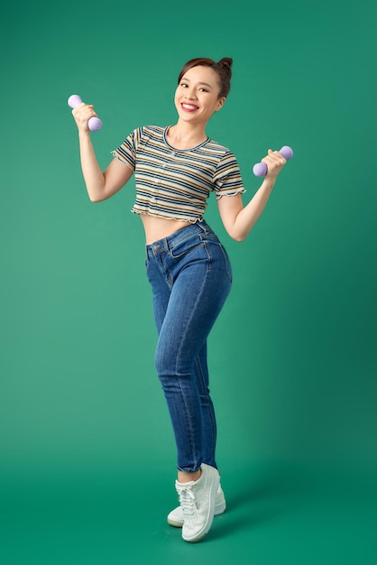 Attractive young Asian woman holding dumbell while standing over green background Full length