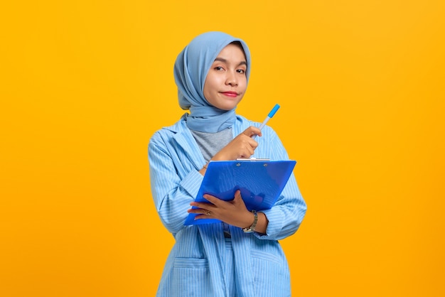 Attractive young Asian woman holding pen and document folder over yellow background