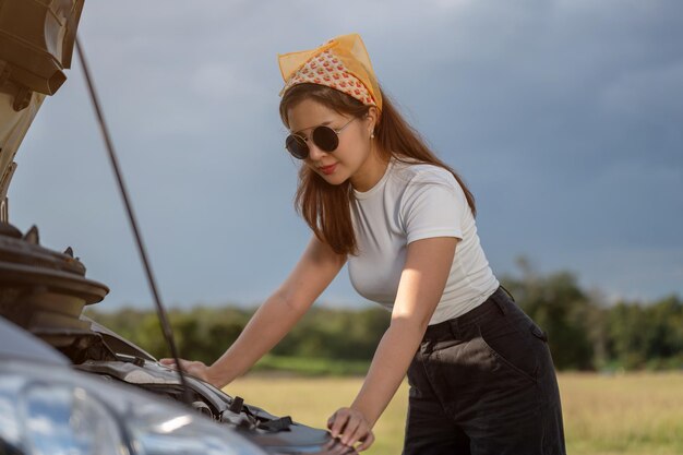 魅力的な若いアジア人女性が田舎への長い旅行の後に車のエンジンをチェックしている