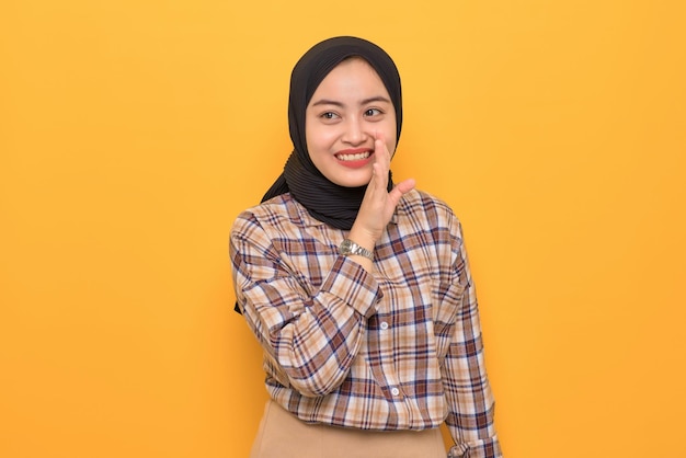 Attractive young Asian woman in plaid shirt hand on mouth telling secret and whispering gossip isolated on yellow background