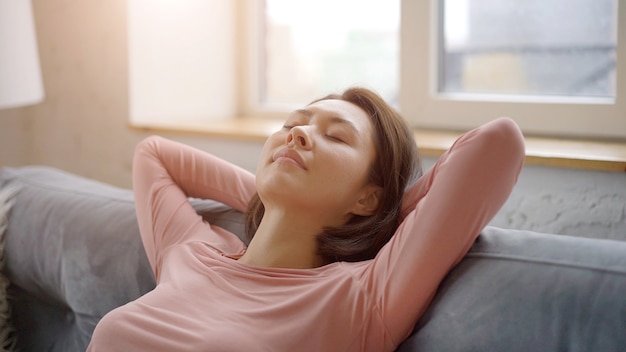 Attractive young asian woman resting on cozy sofa taking deep breath of fresh air holding hands behind head