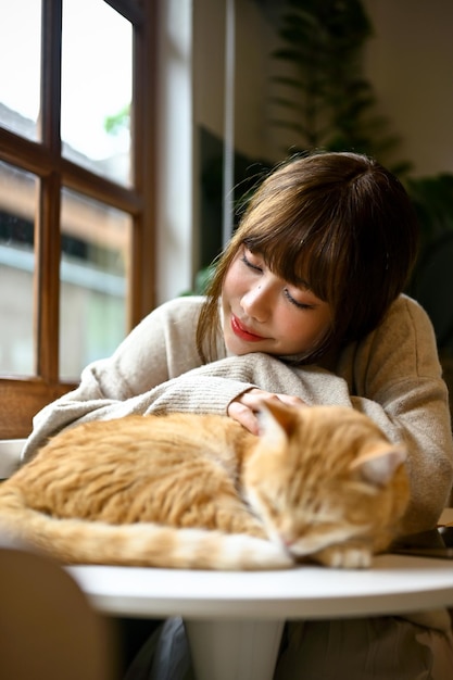 사진 거실에 있는 테이블에서 귀여운 주황색 고양이와 함께 쉬고 있는 매력적인 젊은 아시아 여성
