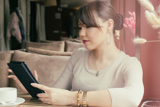 An attractive young asian woman sitting in a cafe is holding a tablet in her hand.