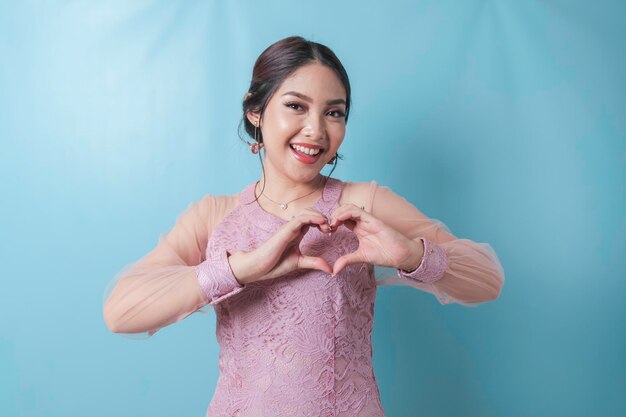 An attractive young Asian woman wearing a modern kebaya feels happy and a romantic shapes heart gesture expresses tender feelings