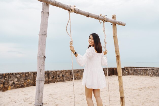 Attractive young asian woman in white dress playing a wooden swing on the beach in summer