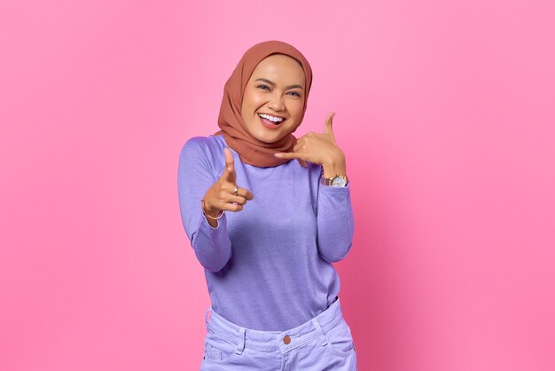 Attractive young Asian woman wink eye pointing to you make call a sign on pink background