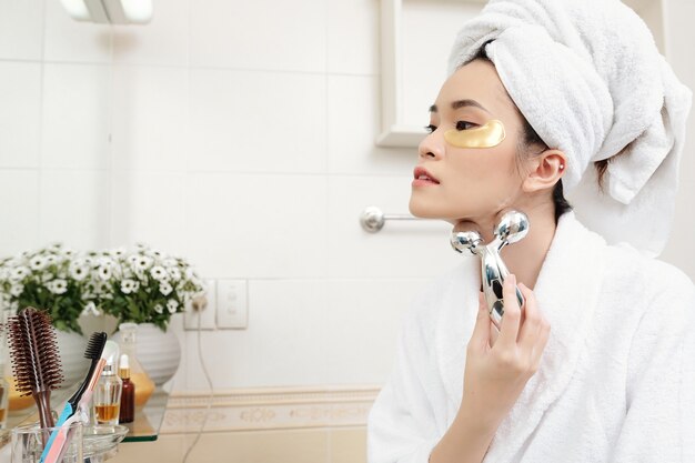 Attractive young Asian woman with undereye patches on her face massaging neck with metal roller to get of wrinkles after taking shower