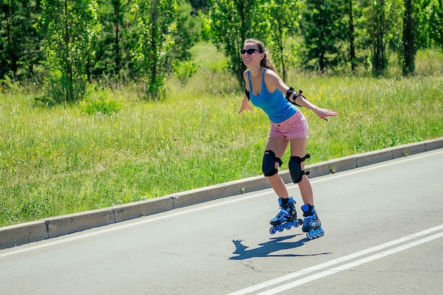 Attractive young athletic slim brunette sexy woman in short
pink shorts and blue top with protection elbow pads and knee pads
ride on roller skatesit in the park