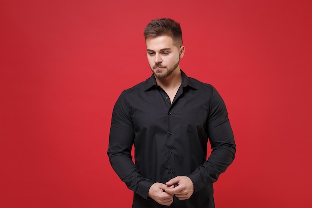 Attractive young bearded guy 20s in classic black shirt posing isolated on bright red wall background studio portrait. People sincere emotions, lifestyle concept. Mock up copy space. Looking down.