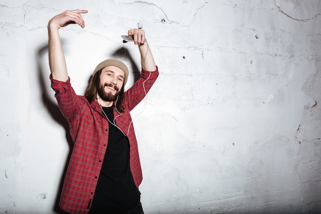 attractive young bearded hipster man wearing hat dressed in shirt in a cage isolated over wall looking at front while listening music with earphones