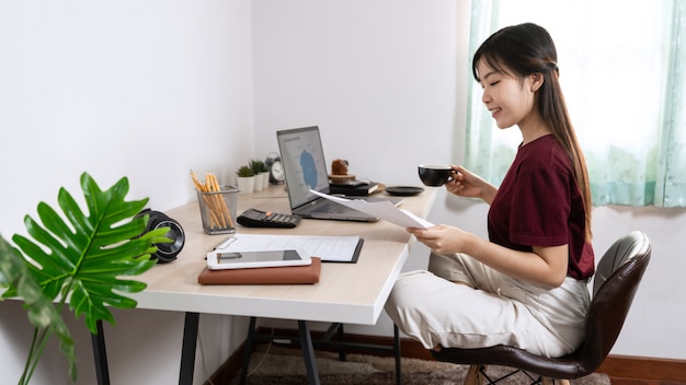 Attractive young beautiful asian woman Entrepreneur or freelancer working at home with laptop business reports and online communications on living room sofa, working remotely access concept.