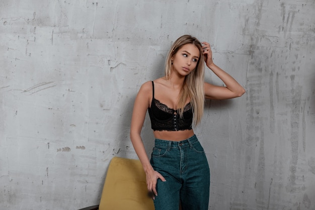 Attractive young beautiful blond woman in stylish blue jeans in a stylish vintage black lace sconce posing indoors near a soft vintage chair. Charming sexy girl.