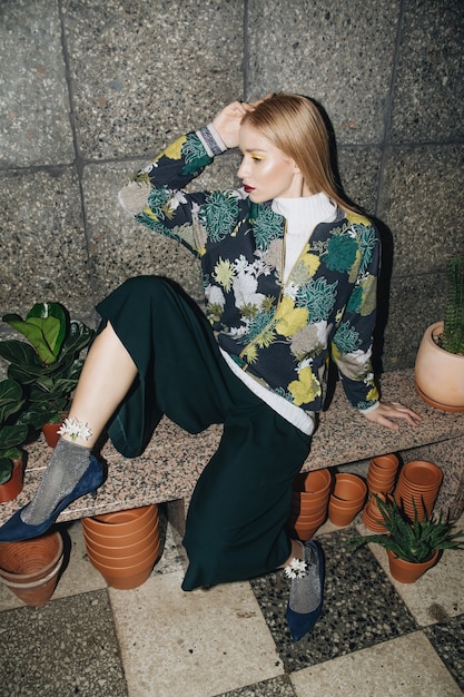 Attractive young blond woman posing in the flower boutique in a stylish dress with tropical print