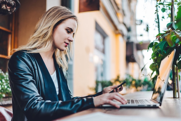 Attractive young blonde business woman dressed in leather jacket spending her lunch time at street cafe terrace and working on her laptop