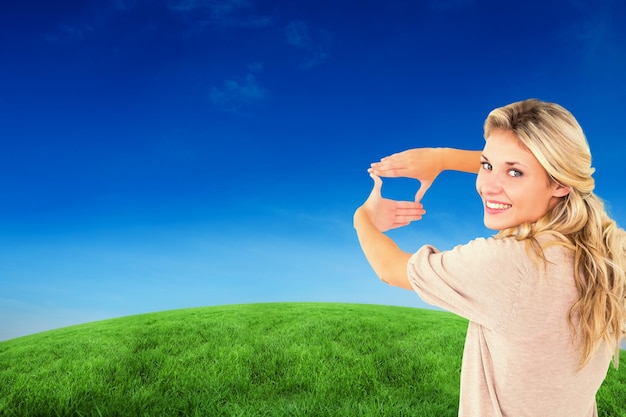 Attractive young blonde framing with her hands against green hill under blue sky