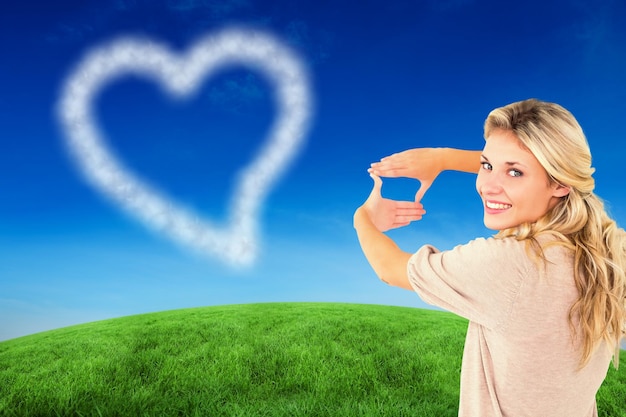 Attractive young blonde framing with her hands against green hill under blue sky