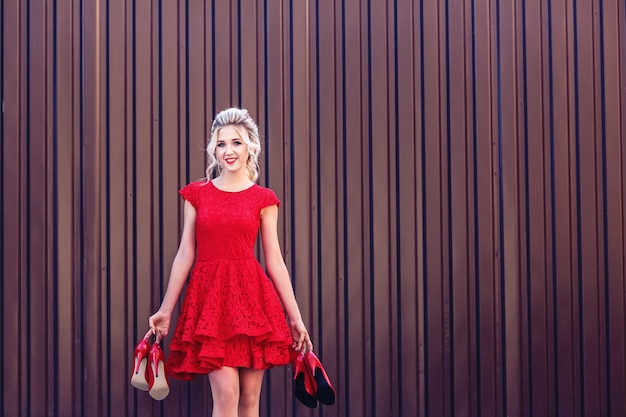 Привлекательная молодая блондинка в красном платье держит в руках много красных туфель. Понятие покупок и продаж