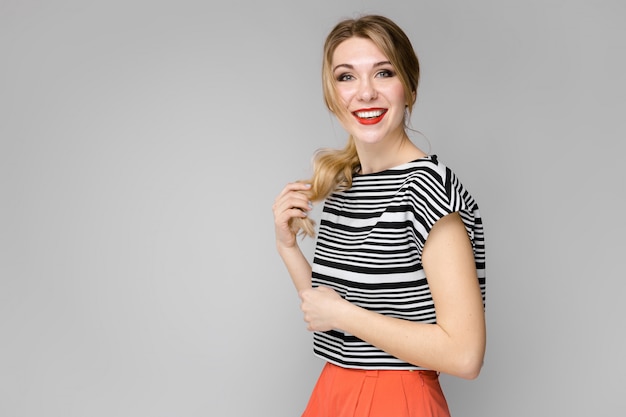 Attractive young blonde girl in striped blouse smiling holding her hair standing on gray wall