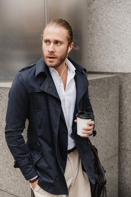 Attractive young blonde haired man in formal wear talking through earphones, holding takeaway coffee cup while standing outdoors on a city street