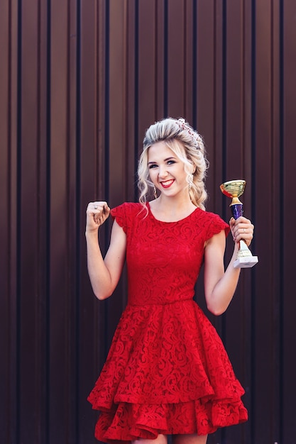 Attractive young blonde in a red dress holding a winner's cup in her hands on a brown background.