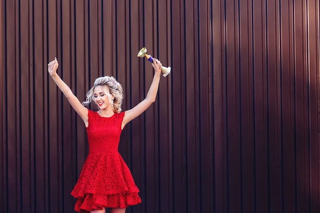 Attractive young blonde in a red dress holds the champion cup and is very happy.