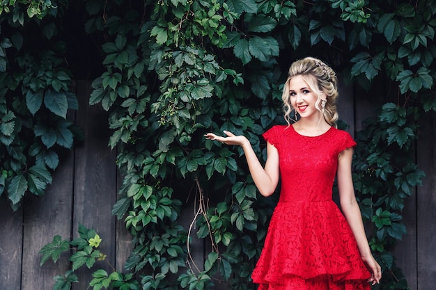 Attractive young blonde in red dress holds copy space on palm against backdrop of wild vineyard.