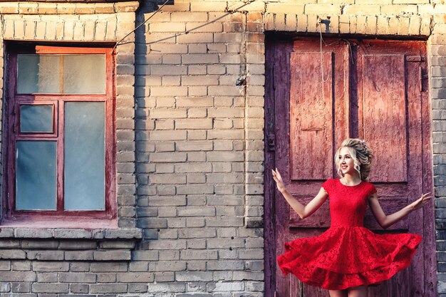 Привлекательная молодая блондинка в красном платье позирует на фоне старого кирпичного здания.