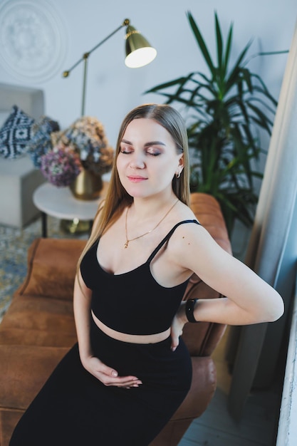 Photo attractive young blonde woman in black top and skirt sitting in brown chair and posing elegant woman in a soft chair