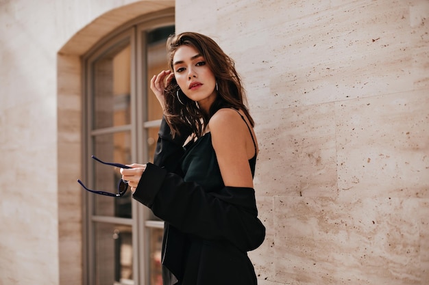 Attractive young brownhaired lady with bright makeup dark dress and black jacket posing outdoors against beige building background and looking into camera