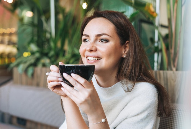 Attractive young brunette smiling woman in white casual dress with cup of coffee in hands in cafe