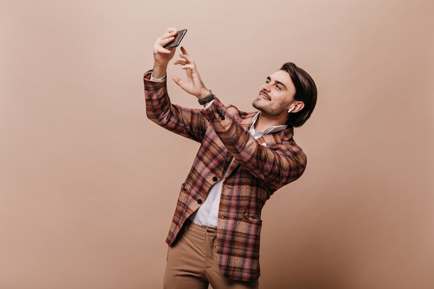 Attractive young brunette in white shirt and brown plaid blazer smiling, video chatting with phone and headphones isolated over beige wall