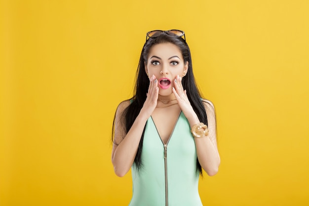 Attractive young brunette woman in a green swimsuit on a yellow. surprised woman in a turquoise bodysuit