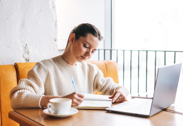 Attractive young brunette woman student freelancer with cup of tea using laptop working in cafe