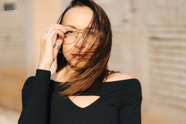 Attractive young brunette woman wearing a black top and transparent glasses posing on the street.