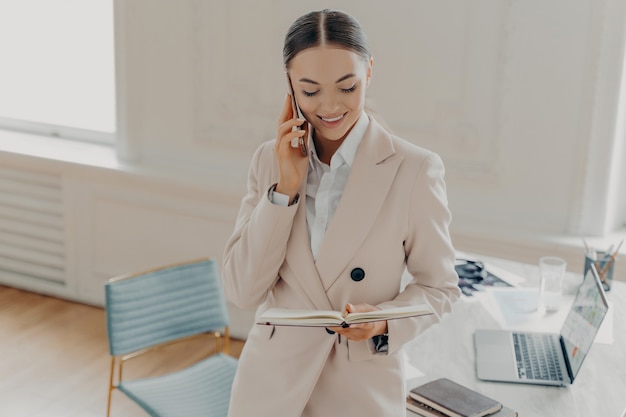 Attractive young business lady talking on mobile phone with boss about working issues and looking at notepad while standing near work desk with laptop in light stylish office