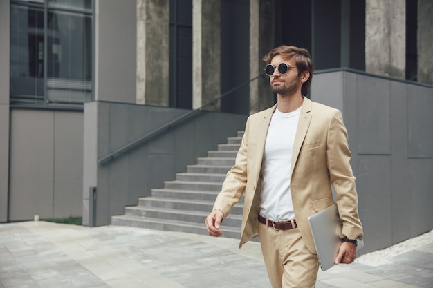 Attractive young businessman in beige suit and trendy sunglasses carrying wireless laptop while walking near office center
