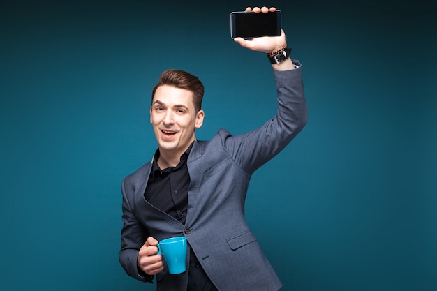 Attractive young businessman in grey jacket and black shirt hold blue cup and show the phone