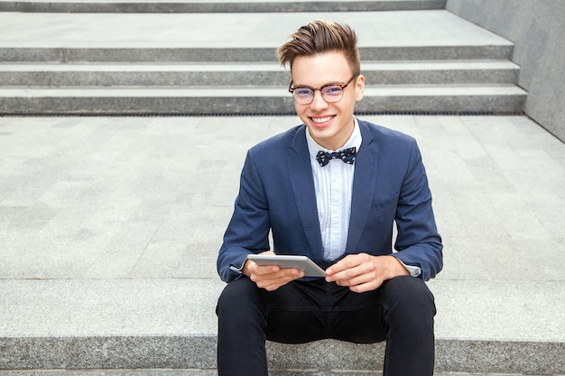 Attractive young businessman sitting with tablet and coffee in hands on office building background. holding tablet and looking at camera with toothy smile.