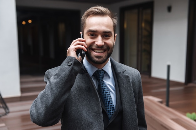 Attractive young businessman wearing suit walking outdoors, using mobile phone, talking