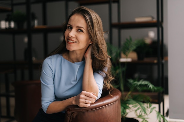 Attractive young businesswoman wearing stylish light blue dress sitting at the chair in office with modern interior, looking at the camera. Portrait of charming business lady.