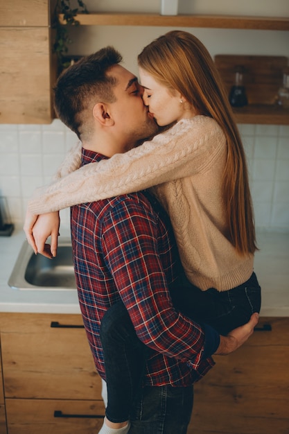 Attractive young caucasian couple kissing in the kitchen in the morning while man is holding his girlfriend on his arms.