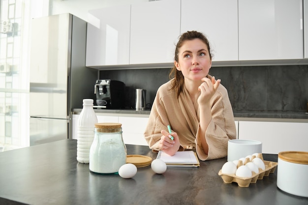 Photo attractive young cheerful girl baking at the kitchen making dough holding recipe book having ideas