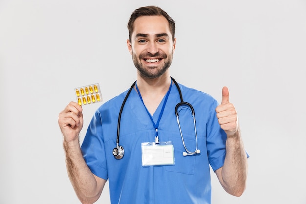 Attractive young cheerful male doctor wearing unifrom standing isolated over white wall, showing pills capsules