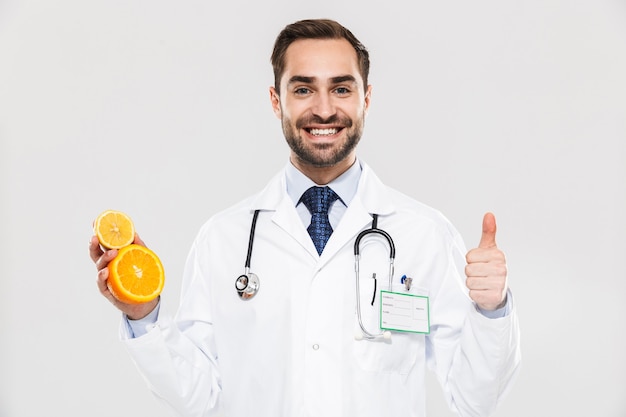 白い壁の上に孤立して立っているunifromを身に着けている魅力的な若い陽気な男性医師、スライスされたオレンジを示しています