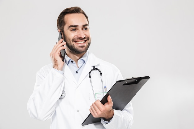 Attractive young cheerful male doctor wearing unifrom standing isolated over white wall, taking notes while talking on mobile phone