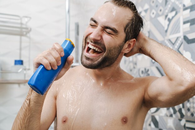 Attractive young cheerful man singing while washing in the shower, holding shampoo bottle